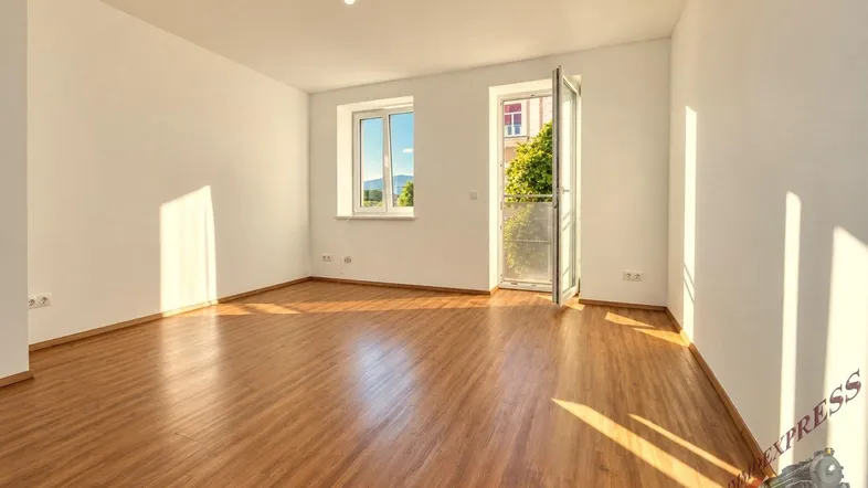 Helles Zimmer mit Holzboden, weißen Wänden und Zugang zu einem französischen Balkon, durchflutet von Sonnenlicht.