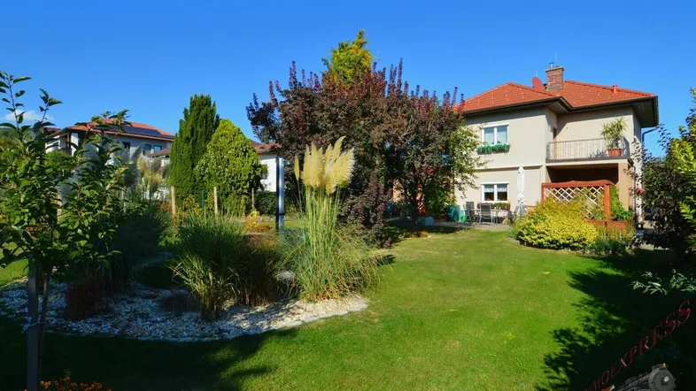 Gepflegter Garten mit Rasenfläche, Zierpflanzen und Blick auf das Haus unter blauem Himmel.