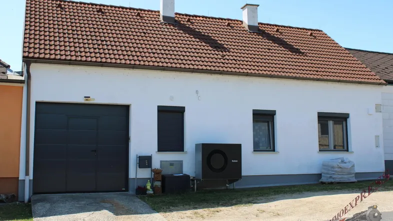 Weißes Einfamilienhaus mit rotem Ziegeldach, Garage und Wärmepumpe im Vorgartenbereich.