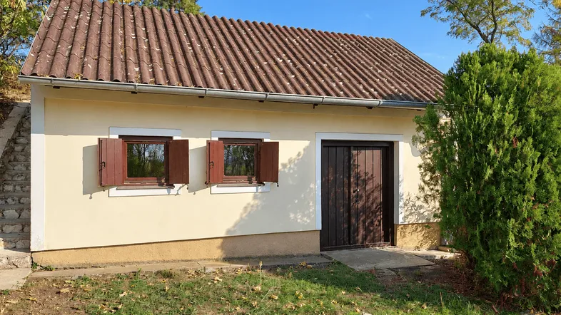 Kleines Gebäude mit Ziegeldach, heller Fassade und braunen Holzfensterläden in ländlicher Umgebung.