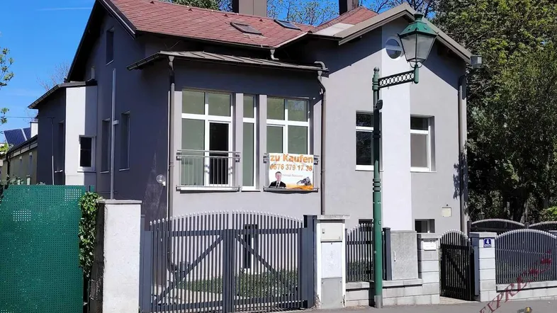 Graue Hausfassade mit weißen Fenstern und einem "Zu Kaufen" Schild an der Vorderseite.