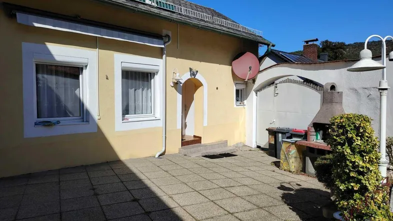 Gepflasterte Terrasse vor dem Haus mit integriertem Grillbereich und Sonnenschutz. Sonnige Außenfläche.