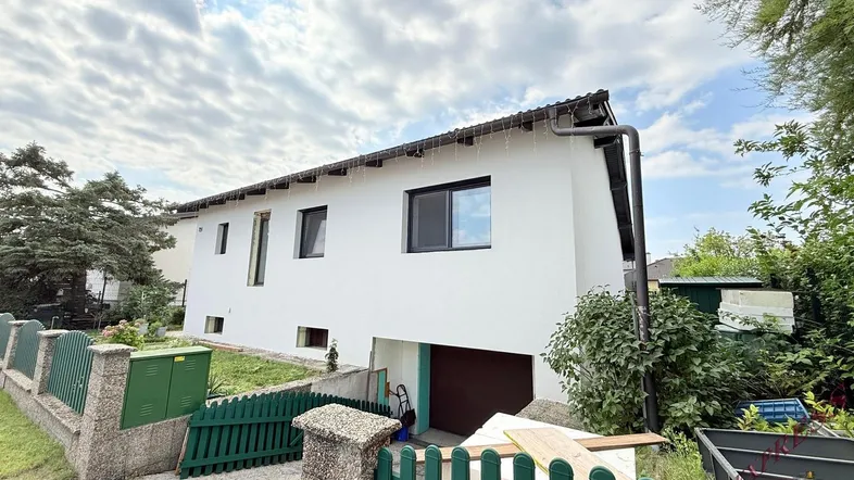 Weißes Einfamilienhaus mit Garage im Untergeschoss und grünem Zaun im Vorgarten.