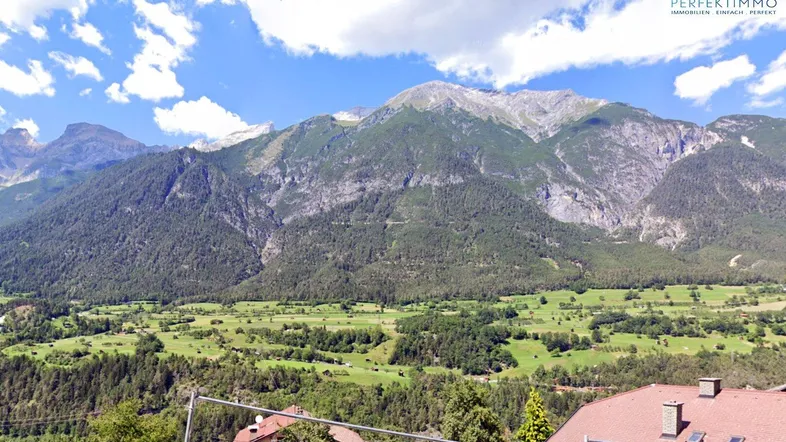 Panoramablick auf majestätische Berge und ein grünes Tal mit vereinzelten Häusern unter blauem Himmel.