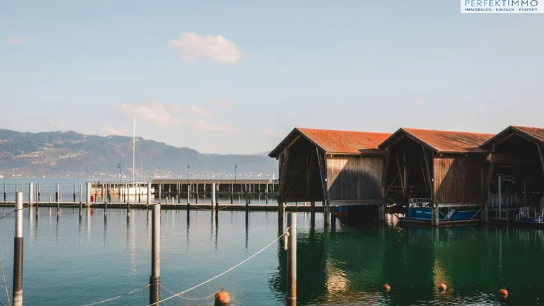 Bootehäuser am See mit Bergen im Hintergrund unter klarem Himmel, umgeben von ruhigem Wasser.
