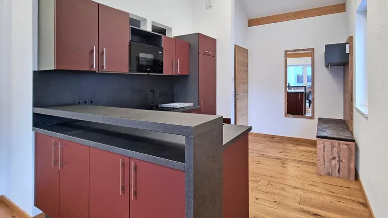 Moderne Küche mit roten Schränken, dunkler Arbeitsplatte und Holzboden, offen zum Eingangsbereich.