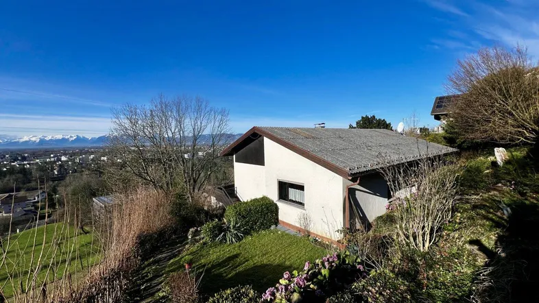Haus auf einem Hügel mit weitem Blick über die Landschaft und schneebedeckte Berge im Hintergrund.