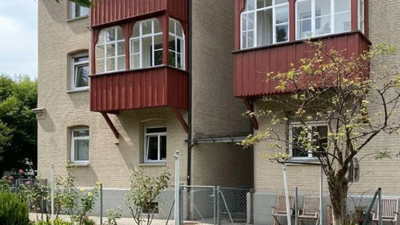 Mehrfamilienhaus mit roter Fassadenverkleidung und mehreren verglasten Balkonen sowie einem kleinen Vorgarten.