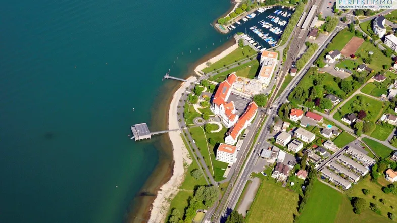 Luftaufnahme einer Küstenlandschaft mit Gebäuden, einem Yachthafen und einem Strandbereich am See.