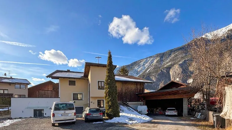 Ländliches Haus mit Bergblick und Parkplätzen im Vordergrund, umgeben von winterlicher Landschaft.