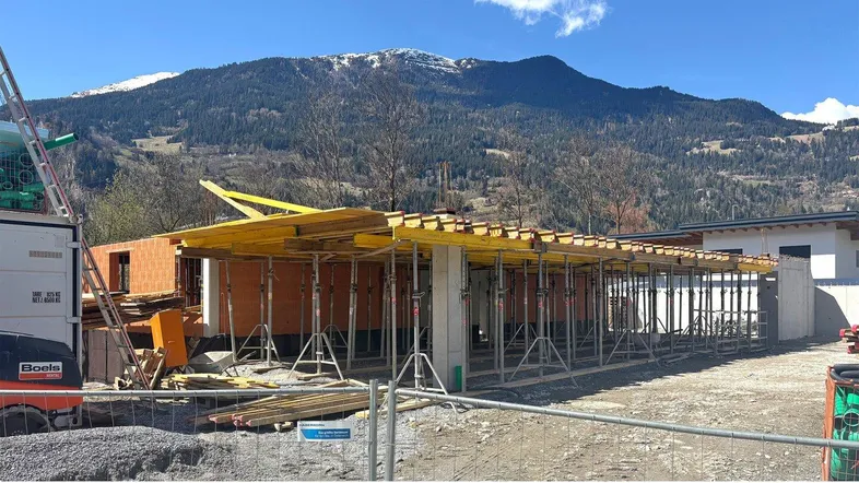Baustelle eines Neubaus mit Gerüst und Baumaterialien vor einer beeindruckenden Bergkulisse unter blauem Himmel.