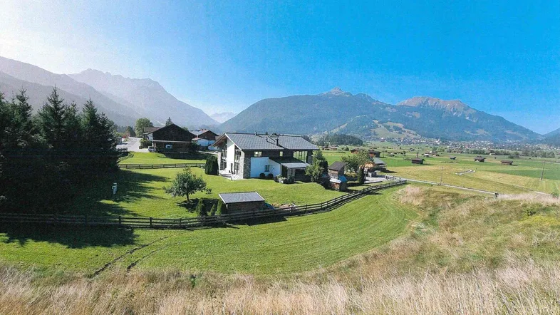 Panoramablick auf das Anwesen mit umliegenden Wiesen und majestätischer Bergkulisse unter blauem Himmel.