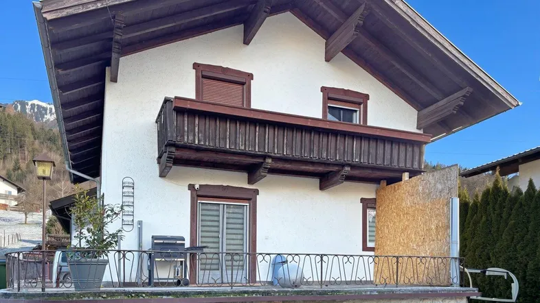Traditionelles Haus mit Balkon und einer Terrasse im Erdgeschoss, umgeben von alpiner Landschaft.