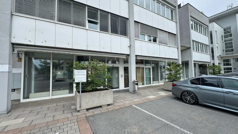 Modernes Geschäftsgebäude mit großen Schaufenstern und Parkplätzen davor unter bewölktem Himmel.