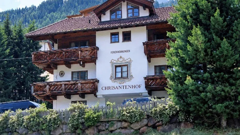 Traditionelles alpines Gebäude mit Holzbalkonen und weißer Fassade, umgeben von grünen Bäumen und Bergen.