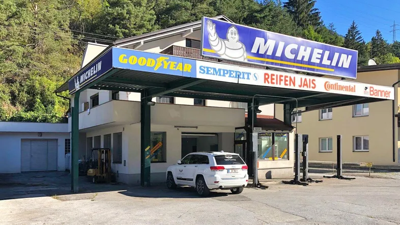 Gebäude mit Reifenservice und Werkstattbereich vor bewaldeter Bergkulisse mit Parkplätzen.