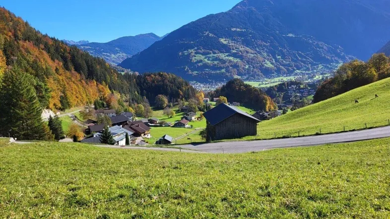 Malerische Berglandschaft mit grünen Wiesen, einem kleinen Dorf und traditionellen Häusern unter blauem Himmel.