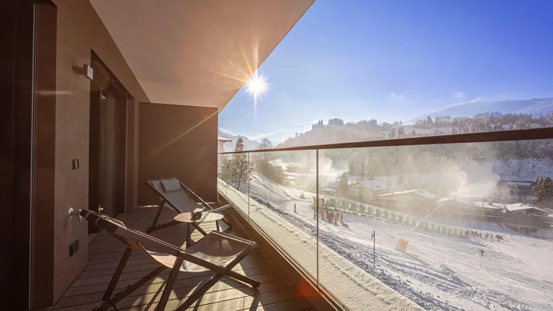 Sonniger Balkon mit Holzboden und Glasbrüstung, bietet weiten Blick auf die verschneite Berglandschaft.