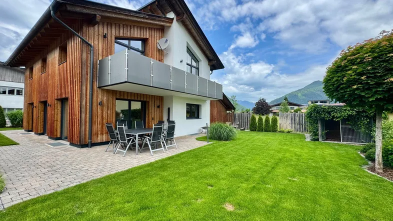 Modernes Haus mit Holzfassade, gepflegtem Garten, Terrasse und Bergblick unter blauem Himmel.