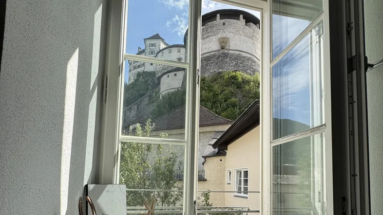 Helles Fenster mit Blick auf eine historische Burg auf einem grünen Hügel unter blauem Himmel.