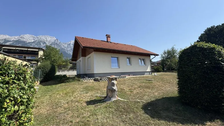 Hausfassade mit Garten und Bergblick unter blauem Himmel an einem sonnigen Tag.