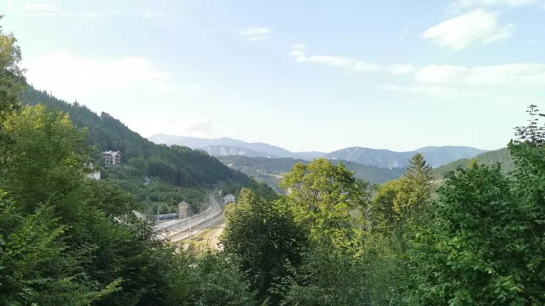 Weitläufiger Blick über ein grünes Tal mit Bergen und einer Eisenbahnlinie im Vordergrund.