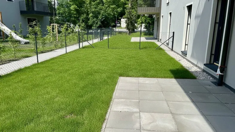 Frisch angelegter Rasenbereich mit gepflasterter Terrasse neben einem modernen Gebäude und Spielplatz.