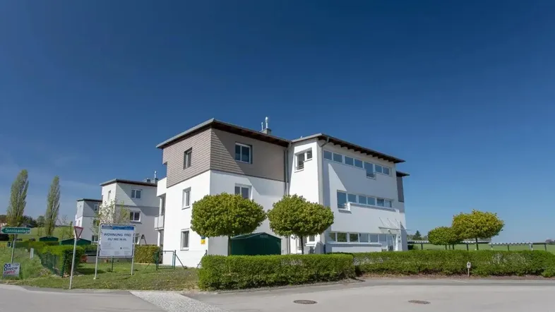 Modernes Mehrfamilienhaus mit weißer Fassade und grüner Umgebung unter blauem Himmel.