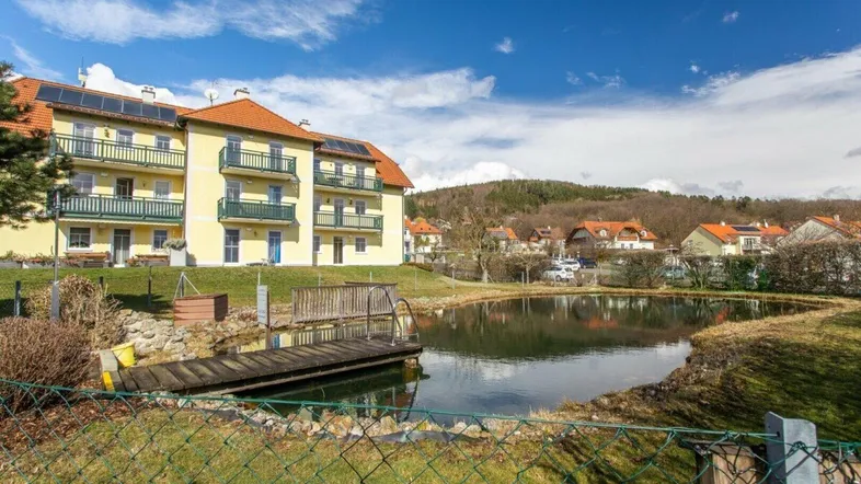 Gelbes Mehrfamilienhaus mit Balkonen und Solaranlage, umgeben von einem Teich und grüner Landschaft.