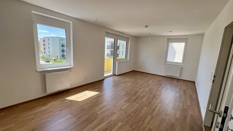 Helles Wohnzimmer mit Laminatboden, weißen Wänden und Zugang zu einem Balkon.