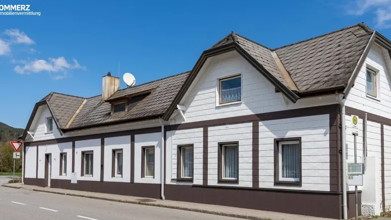 Ansicht eines traditionellen Gebäudes mit weißer Fassade, braunen Akzenten und Satteldach unter blauem Himmel.