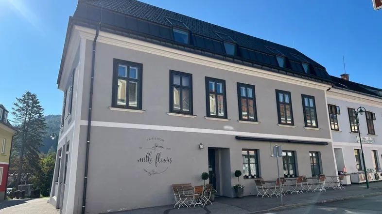 Graue Fassade eines Mehrfamilienhauses mit schwarzen Fenstern und einem Café im Erdgeschoss.