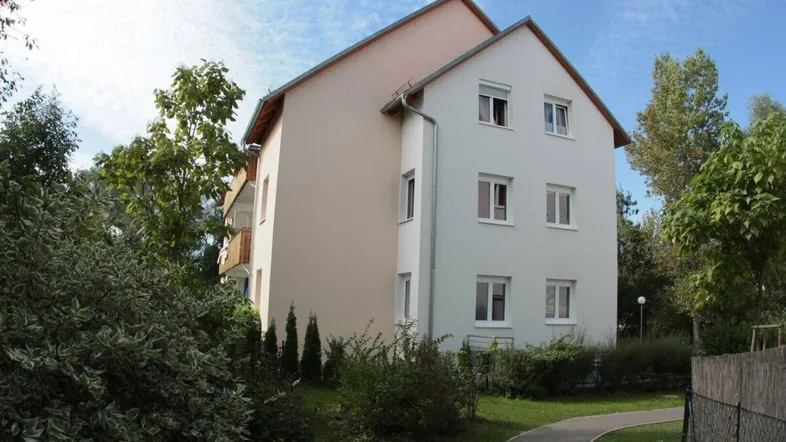 Wohngebäude mit heller Fassade und Satteldach, umgeben von Bäumen und einem Gehweg.