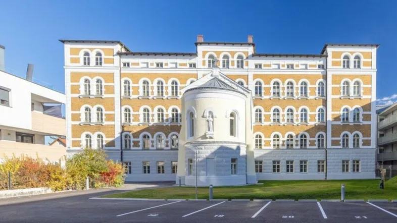 Historisches Gebäude mit markanter Architektur und einem Vorplatz mit Parkplätzen unter blauem Himmel.