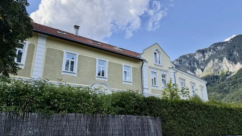 Gelbe Fassade eines historischen Gebäudes mit roten Ziegeldächern und Bergblick unter bewölktem Himmel.