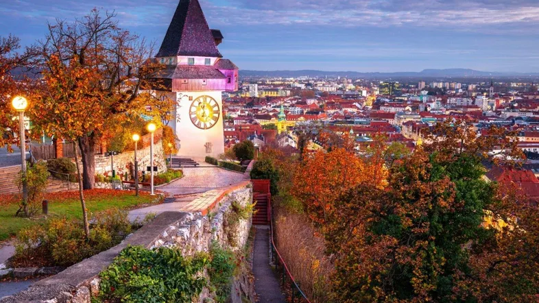 Abendlicher Panoramablick über Graz mit dem beleuchteten Uhrturm und herbstlicher Vegetation.
