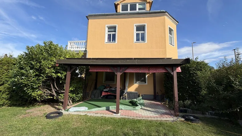 Gelbes Haus mit überdachter Terrasse und Gartenbereich unter blauem Himmel.