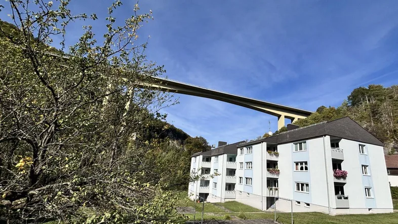 Mehrfamilienhaus in bergiger Umgebung mit einer großen Brücke im Hintergrund unter blauem Himmel.