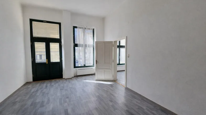 Heller, leerer Wohnraum mit grauem Laminatboden, großen Fenstern und weiß gestrichenen Wänden.