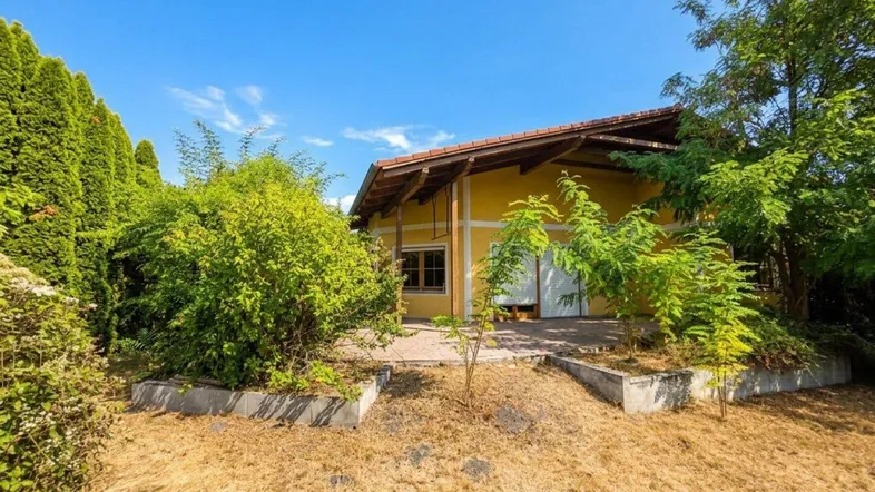 Gelbes Haus mit rotem Dach, umgeben von üppigem Grün und einem sonnigen Gartenbereich.