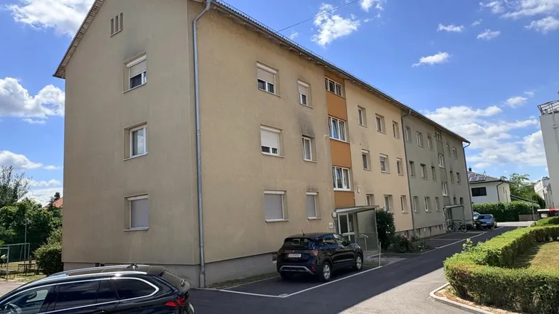 Mehrfamilienhaus mit mehreren Etagen und Parkplätzen vor dem Gebäude unter blauem Himmel.