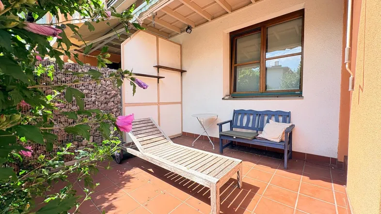 Gemütliche Terrasse mit Liegestuhl und Sitzbank, umgeben von grünen Pflanzen und blühenden Sträuchern.