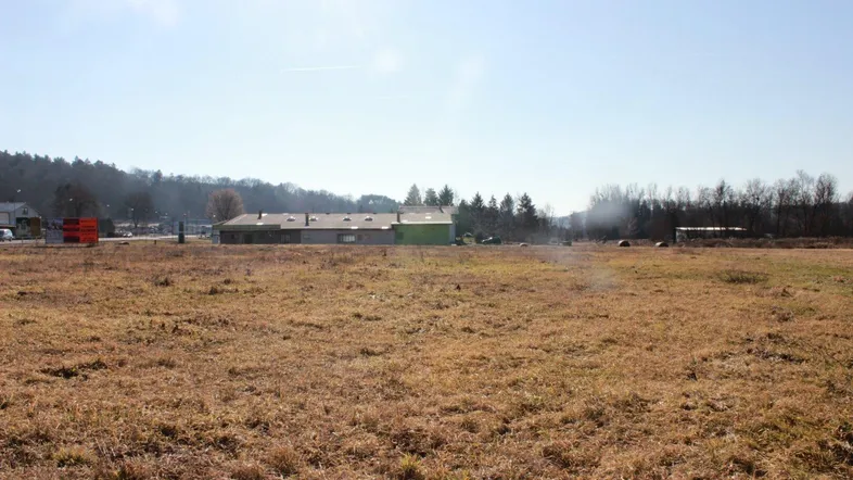 Weitläufiges Feld mit Blick auf entfernte Gebäude und bewaldete Hügel unter blauem Himmel.