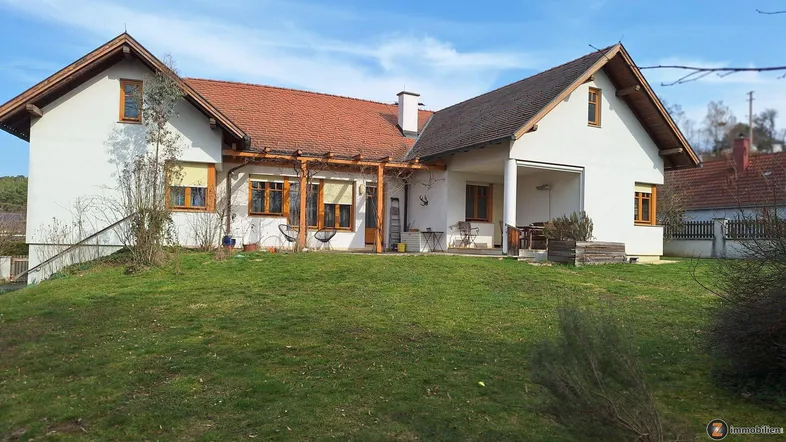 Weißes Einfamilienhaus mit rotem Ziegeldach, großem Garten und überdachtem Terrassenbereich unter blauem Himmel.