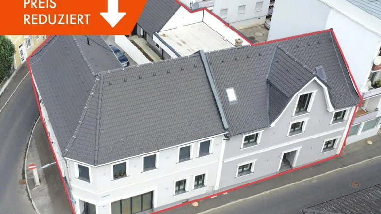 Luftaufnahme eines modernen Eckgebäudes mit hellgrauer Fassade und dunklem Ziegeldach an einer Straße.