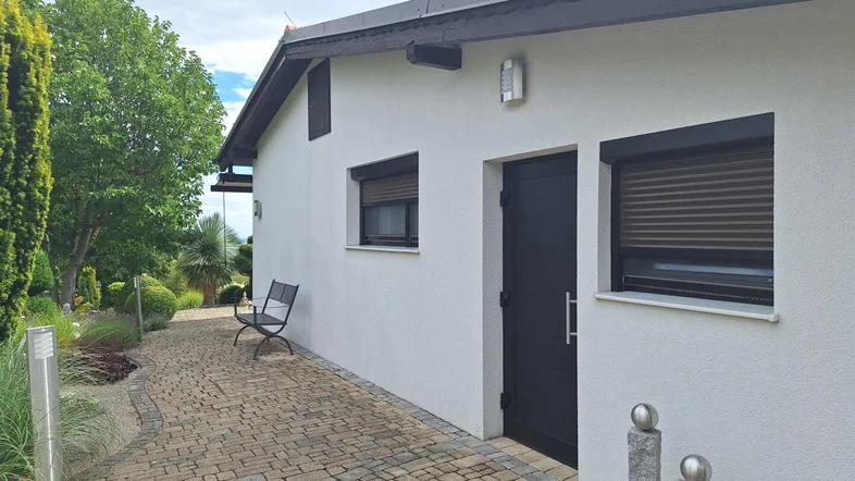 Seitenansicht des Hauses mit gepflastertem Weg, kleinen Fenstern und Rollläden.