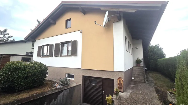 Seitenansicht des Hauses mit Garagenzufahrt und Außentreppe, umgeben von Bepflanzung.