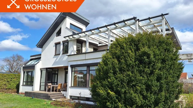 Modernes Haus mit Terrasse, Pergola und gepflegtem Vorgarten unter blauem Himmel.