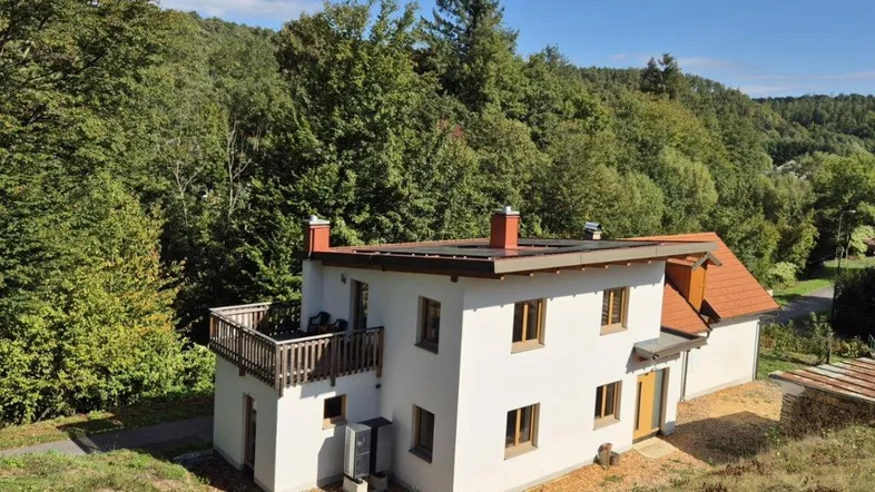 Freistehendes Haus mit weißer Fassade, Flachdach und kleinem Balkon, umgeben von Wald und Natur.