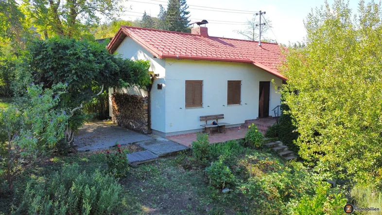 Kleines Einfamilienhaus mit rotem Ziegeldach und Holzstapel an der Seite, umgeben von viel Grün.
